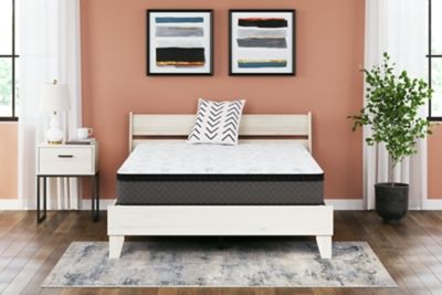 A modern bedroom featuring a white platform bed with a Sierra Sleep® by Ashley 12 Inch Pocketed Hybrid mattress, flanked by a small white nightstand. Above the bed, three horizontal abstract paintings hang on a peach-colored wall.