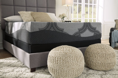 A modern bedroom featuring an Ashley Sleep® 1100 Series Hybrid Mattress on an upholstered gray bed with patterned bedding, flanked by a light-filled window and a bedside table with flowers, accompanied by two woven ottomans.