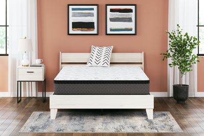 A modern bedroom featuring a white wooden bed frame with a Sierra Sleep® by Ashley 12 Inch Pocketed Hybrid mattress. There are three abstract paintings above the bed, a white nightstand with a lamp, and a potted plant.