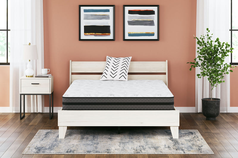 A modern bedroom featuring a white wooden bed with a Sierra Sleep® by Ashley 10 Inch Pocketed Hybrid Mattress and a striped pillow, flanked by a nightstand and a tall floor lamp, against a salmon-colored wall adorned with abstract